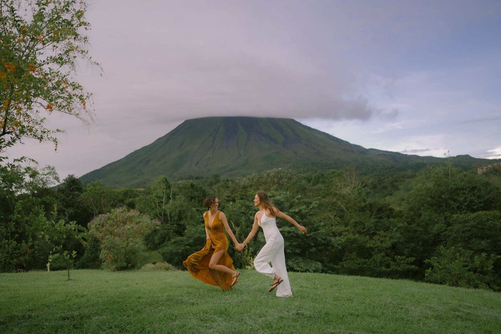 Two people running hand in hand across a green lawn with a volcano behind them, Costa Rica