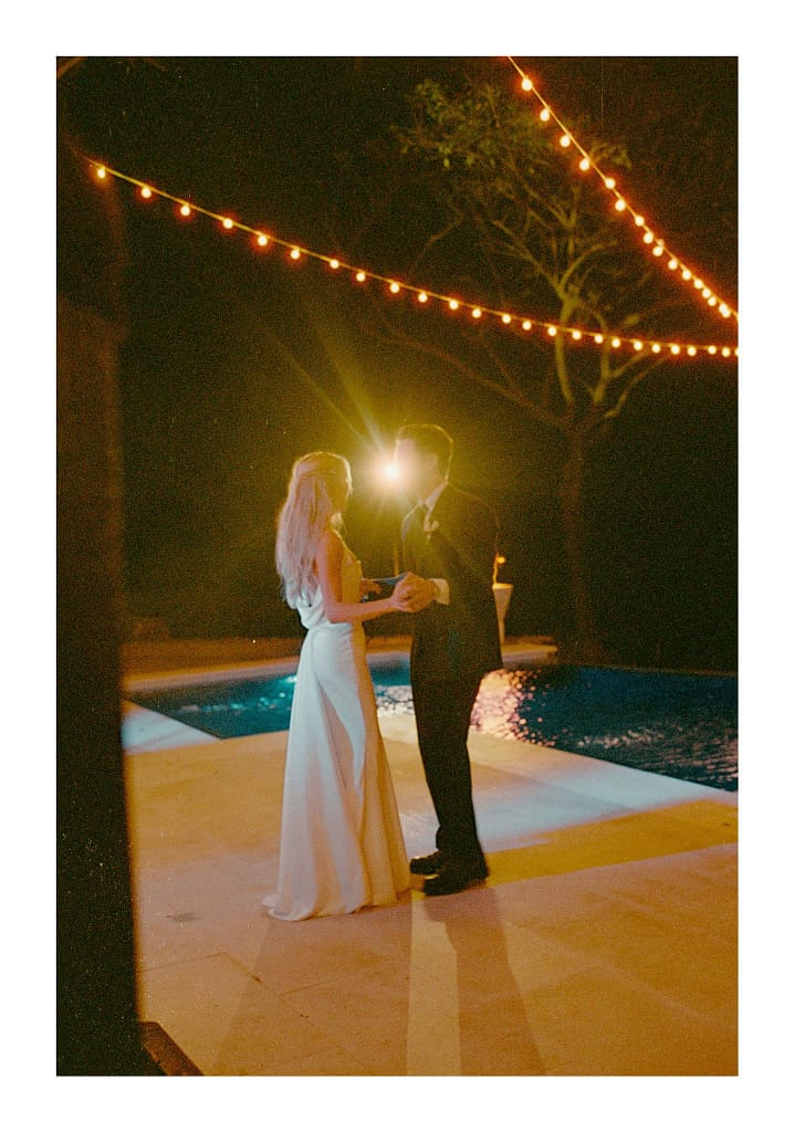 Film photograph of a couple dancing under string lights beside a pool, glowing from a light behind them