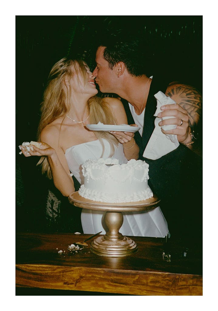 Film photograph of a couple sharing a kiss behind a white wedding cake on a gold stand