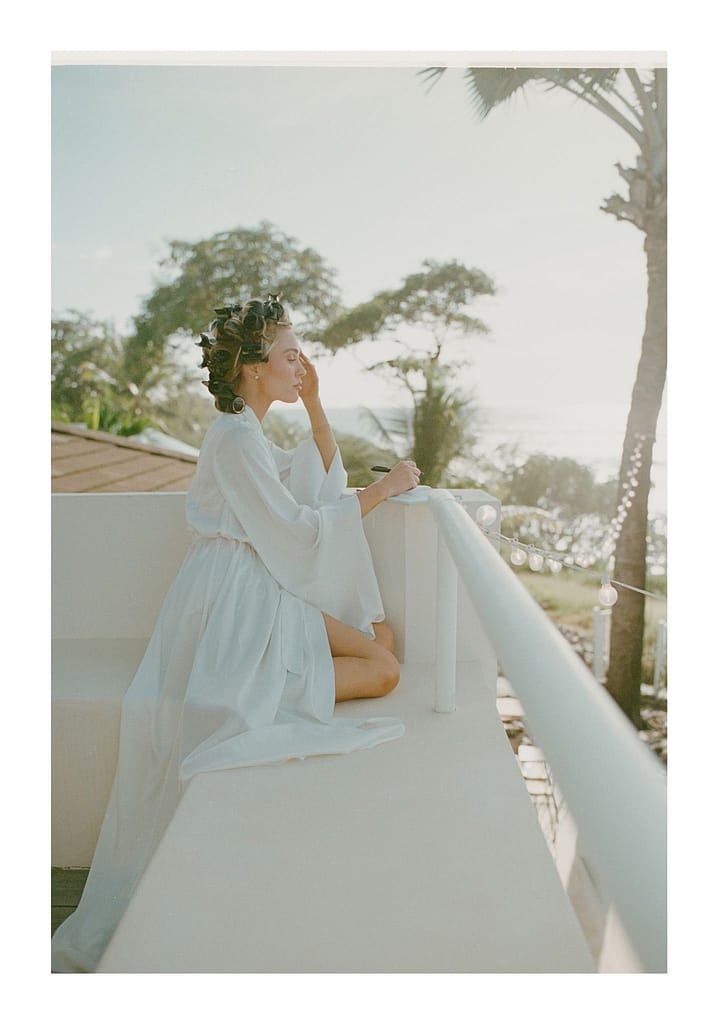 Film photograph of a bride in a white robe sitting on a balcony rail with palm trees behind her
