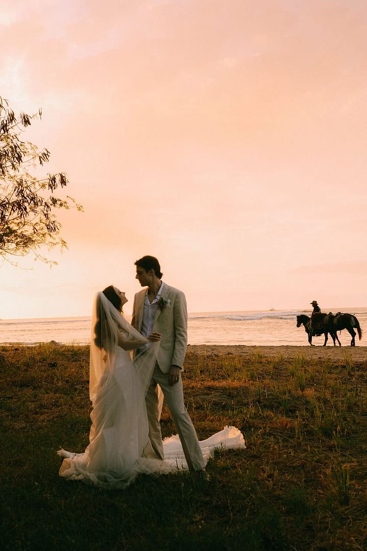 Bride and groom at sunset on a Tamarindo beach as a horse rider passes in the background, Costa Rica
