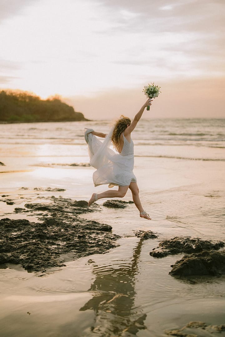 Bride leaping with her bouquet raised on a beach at sunset, Costa Rica