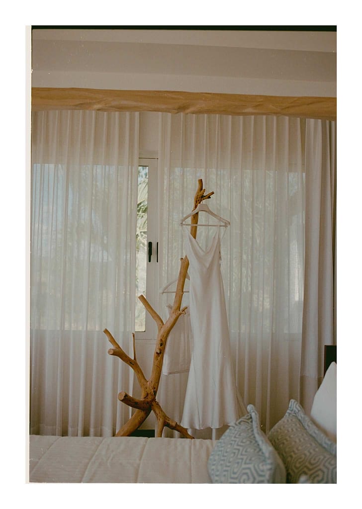 Film photograph of a wedding dress hanging on a driftwood branch beside sheer curtains