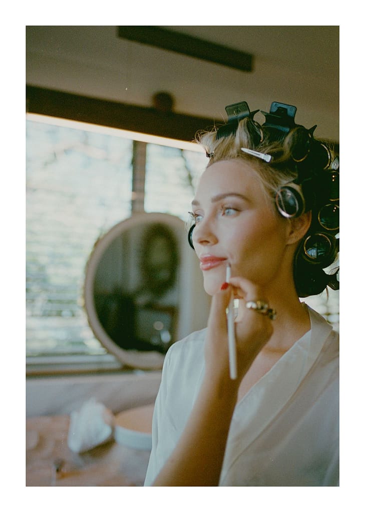 Film portrait of a bride in hair rollers touching up her makeup by a window