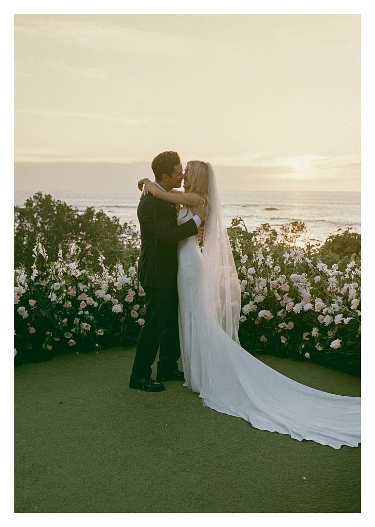 Film photograph of a couple embracing at sunset above the ocean, the bride’s long veil behind her and white flowers around them