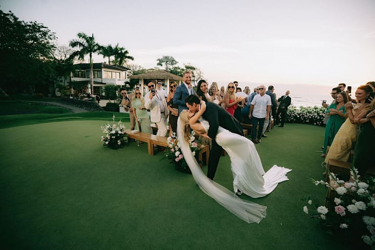 Bride dipped for a kiss in the aisle as guests cheer at a sunset wedding ceremony on the lawn, Costa Rica