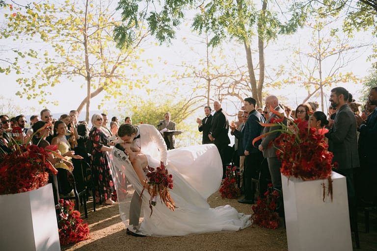 Bride leaning in to greet a guest at the end of a ceremony aisle lined with red flowers, Costa Rica wedding